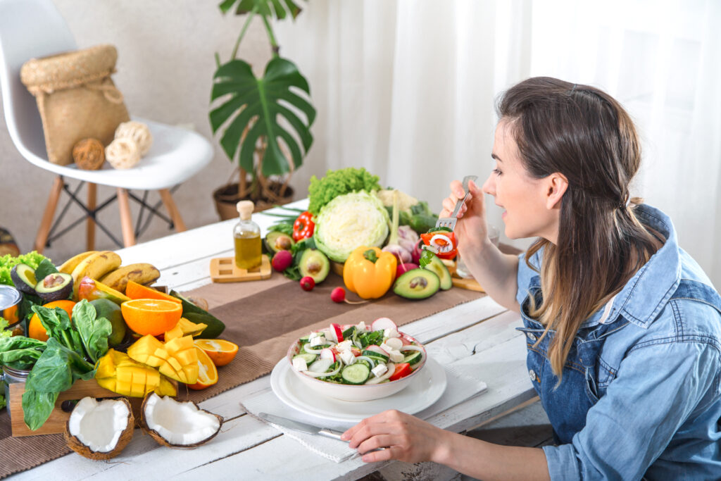 Mulher comendo uma salada fresca enquanto aprecia uma refeição saudável, com frutas, vegetais e óleos vegetais dispostos sobre a mesa em um ambiente iluminado e acolhedor.
