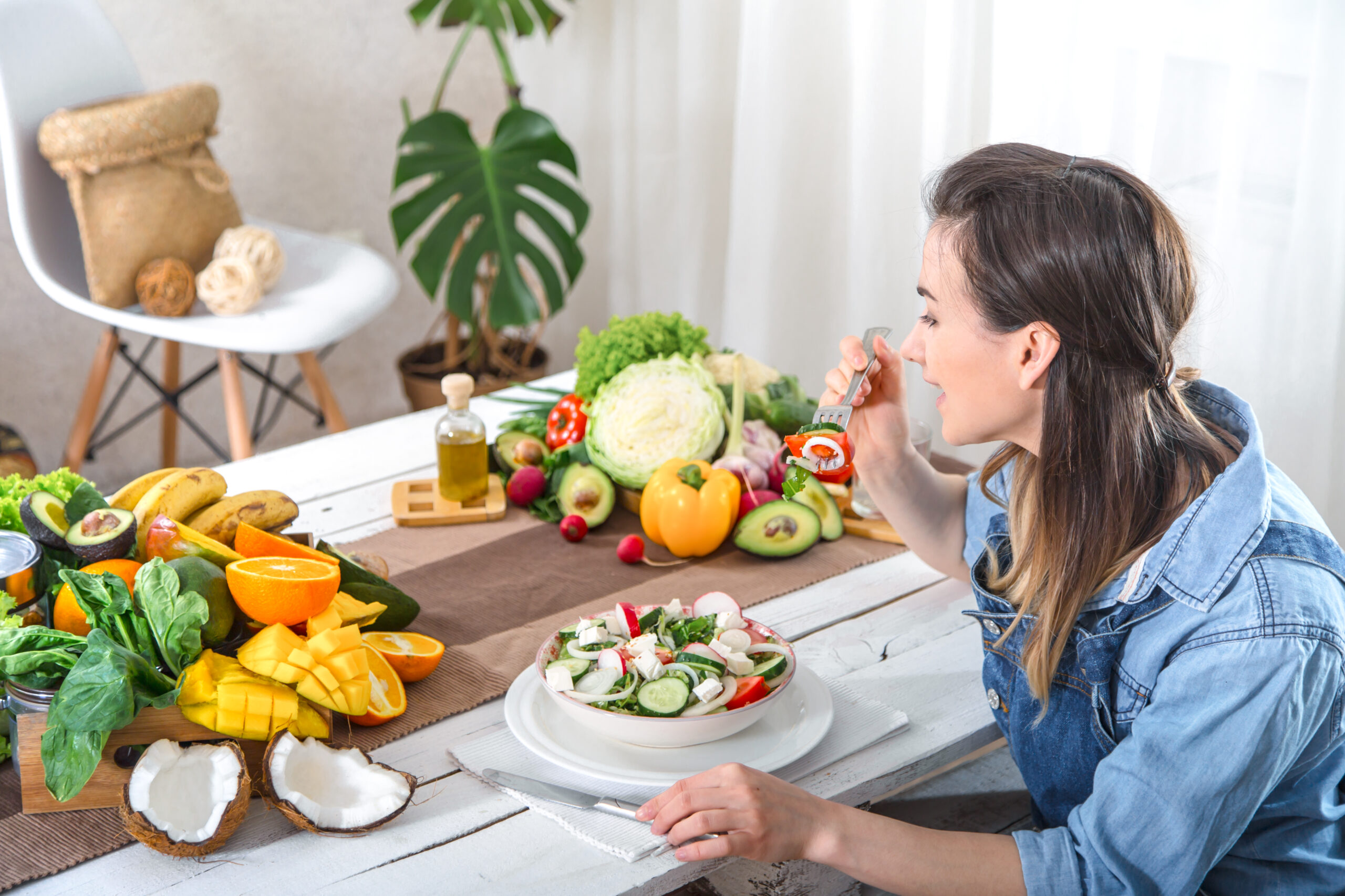 Mulher comendo uma salada fresca enquanto aprecia uma refeição saudável, com frutas, vegetais e óleos vegetais dispostos sobre a mesa em um ambiente iluminado e acolhedor.