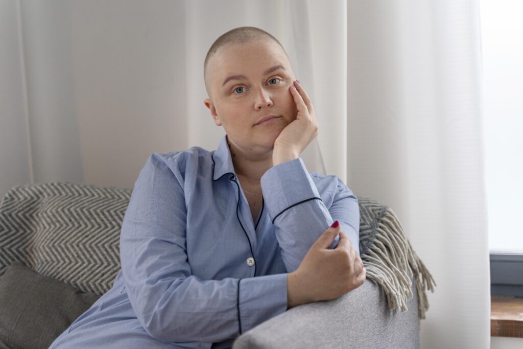 Uma mulher com a cabeça raspada, sentada no sofá, apoiando o rosto na mão com uma expressão pensativa. Ela parece calma, mas reflexiva, vestindo uma camisa azul clara."