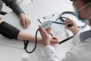 Profissional de saúde realizando medição de pressão arterial em paciente durante consulta médica, com estetoscópio e documentos na mesa.