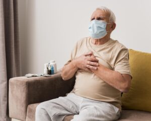 A imagem mostra um homem idoso com uma máscara facial, sentado em um sofá, segurando o peito com uma expressão de desconforto, indicando possível dor no peito. Ao lado dele, há uma mesa com diversos frascos de medicamentos e outros itens relacionados ao tratamento. O ambiente parece ser o interior de uma casa, com móveis simples e aconchegante
