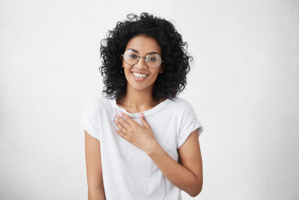 Mulher sorrindo, com cabelos cacheados e óculos, usando uma camiseta branca.