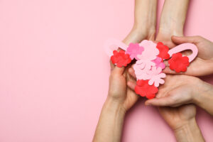 
A imagem mostra uma representação estilizada do sistema reprodutor feminino, formada por peças de papel colorido em tons de rosa e vermelho, organizadas para criar uma ilustração do útero. As peças em forma de flores foram dispostas de forma cuidadosa e são seguradas por mãos de diferentes pessoas, sugerindo cuidado e atenção à saúde feminina. O fundo é rosa, o que cria uma sensação suave e acolhedora.