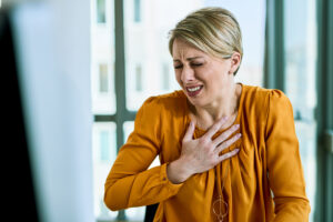 Mulher com expressão de dor no peito, visivelmente aflita, em um ambiente de escritório, sugerindo um possível sintoma de infarto.