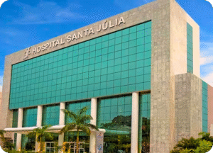 Fachada externa moderna e imponente do Hospital Santa Júlia em Manaus. O edifício é alto, com revestimento em mármore cinza-claro e grandes janelas espelhadas em tom esverdeado/azul. O nome "HOSPITAL SANTA JÚLIA" está em destaque no topo da estrutura. Há palmeiras e um céu azul ao fundo.