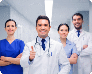 
Uma equipe de quatro profissionais de saúde, composta por dois homens e duas mulheres, está em pé em um corredor de hospital. Todos estão sorrindo e com os braços cruzados, exceto o médico no centro, que está em primeiro plano e dando um "joinha" com a mão direita. Ele usa um jaleco branco e um estetoscópio. A equipe transmite confiança e profissionalismo.
