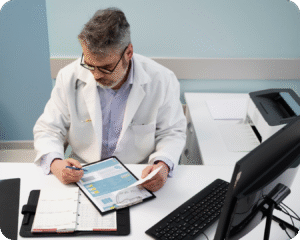  Um médico, com jaleco branco e óculos, está sentado em sua mesa, analisando documentos e resultados de exames. Ele parece concentrado e atencioso. Há um estetoscópio em volta do pescoço.
