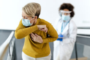 A imagem mostra uma mulher loira, de meia-idade, vestindo uma blusa de lã mostarda, com a mão direita pressionando o peito. Ela está de pé, com uma expressão de desconforto ou dor. Ao fundo, fora de foco, um médico com máscara e escudo facial a observa. Ambos parecem estar em um corredor de hospital. A mulher está usando máscara cirúrgica, assim como o médico.