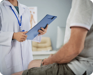 Uma médica de jaleco branco está segurando uma prancheta azul e conversando com um paciente que está sentado em uma maca de hospital.