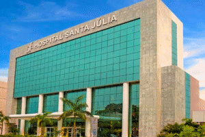  A imagem mostra a fachada de um prédio moderno e grande, com a entrada principal em destaque. As paredes do edifício são de pedra ou mármore cinza-claro, e as janelas são de vidro azul-esverdeado. O nome HOSPITAL SANTA JÚLIA está escrito em letras grandes e cinza no topo da fachada. Em frente ao prédio, há palmeiras e outras plantas, e o céu está parcialmente nublado.