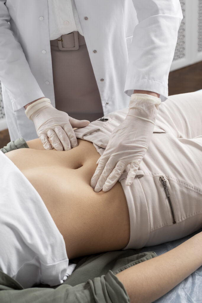 Um médico com jaleco branco e luvas descartáveis examina o abdômen de um paciente deitado, pressionando suavemente a região com as duas mãos para diagnosticar a causa de uma dor.