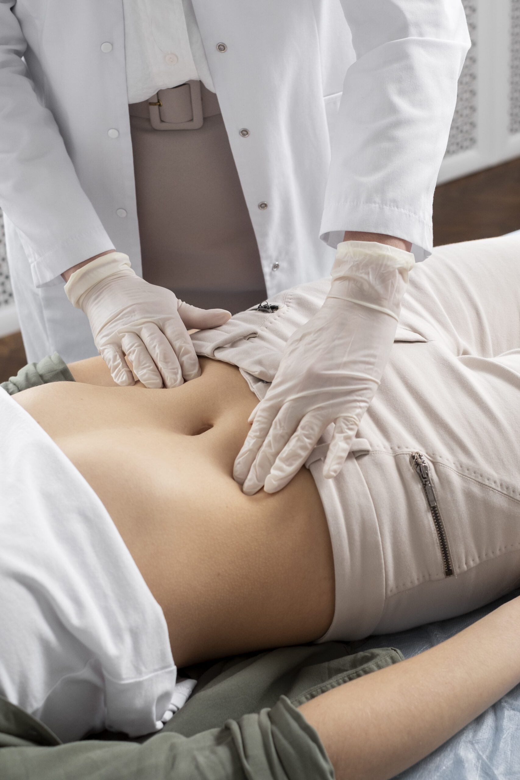 Um médico com jaleco branco e luvas descartáveis examina o abdômen de um paciente deitado, pressionando suavemente a região com as duas mãos para diagnosticar a causa de uma dor.
