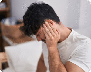 Um homem jovem, com cabelo escuro, está sentado em um ambiente que parece ser um quarto. Ele está com a mão direita na orelha, com uma expressão de desconforto ou dor, como se estivesse com tontura ou dor de cabeça.