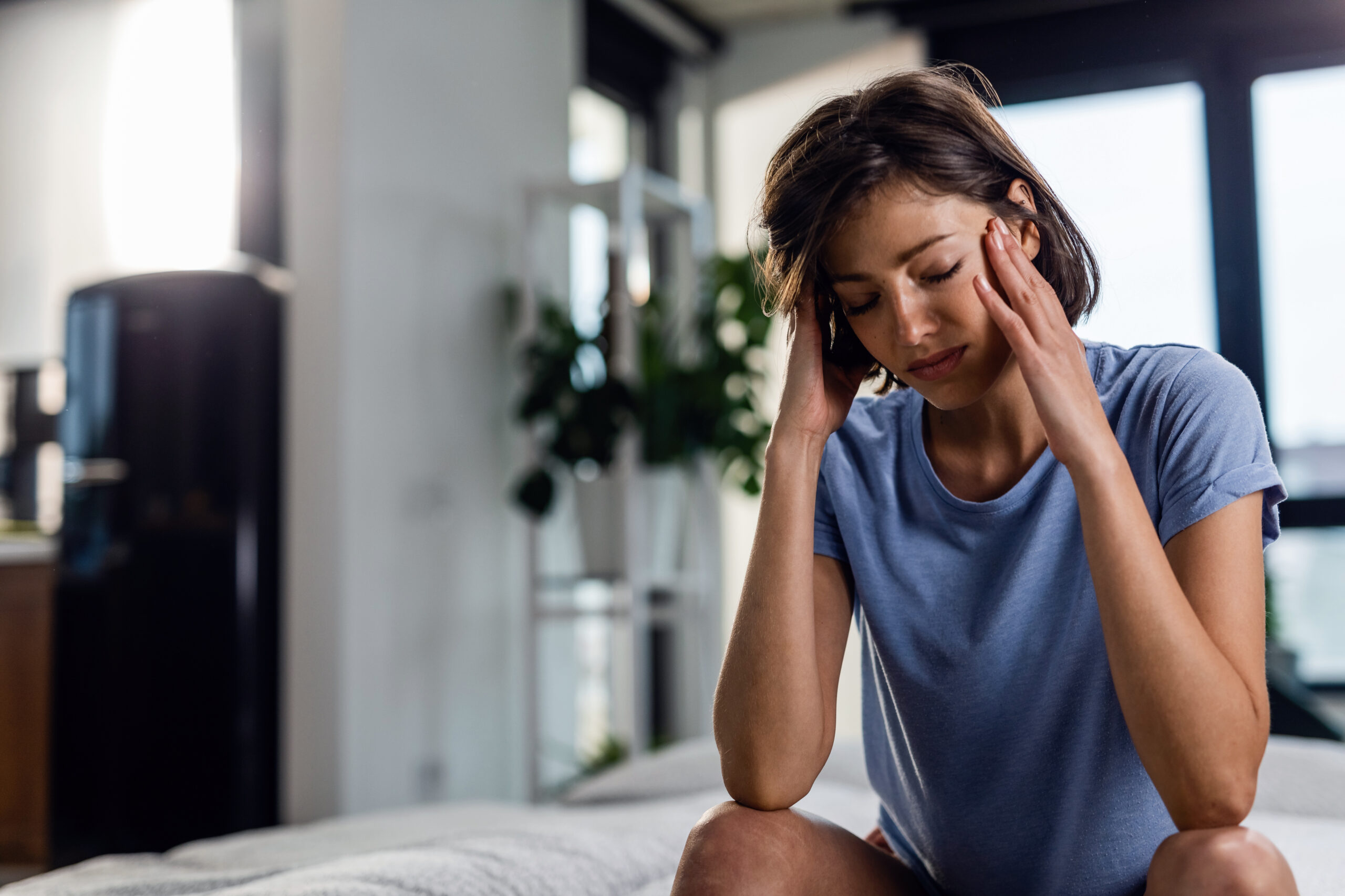 Uma mulher com cabelo curto e castanho, vestindo uma camiseta azul, senta-se na cama. Ela massageia as têmporas com as duas mãos, com os olhos fechados e uma expressão de desconforto ou dor de cabeça.