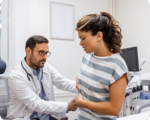 Um médico de jaleco branco está examinando uma paciente com dor abdominal. Ele está sentado ao lado dela e toca sua barriga para fazer o diagnóstico, enquanto ela se inclina para a frente e segura a região da dor.