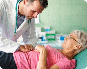 Um médico, com jaleco branco e estetoscópio, examina uma paciente idosa, deitada em uma maca. Ele segura a mão dela com ambas as mãos, demonstrando atenção e cuidado. A paciente, que usa óculos, olha para ele.