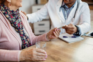 Uma médica ou enfermeira, usando jaleco branco, senta-se em uma mesa de madeira. Ela conversa com uma paciente de idade, que usa um cachecol florido e um casaco rosa. O profissional de saúde segura um frasco de remédio e estende-o para a paciente. Sobre a mesa, há um copo com água, uma caneta e um caderno.