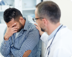  Um médico, de jaleco branco, coloca a mão no ombro de um paciente para confortá-lo. O paciente, um homem, está sentado e com a mão no rosto, como se estivesse com dor ou tontura.