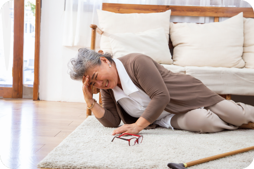 Mulher idosa, deitada no chão ao lado de um sofá e de sua bengala, com a mão na cabeça em uma expressão de dor ou mal-estar. A cena sugere uma queda súbita e incapacitante, possivelmente devido a um AVC.
