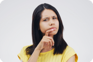 Mulher jovem, com cabelos escuros e vestindo uma blusa amarela, olhando diretamente para a câmera com uma expressão séria e confusa, com a mão no queixo em um gesto pensativo ou de incerteza.