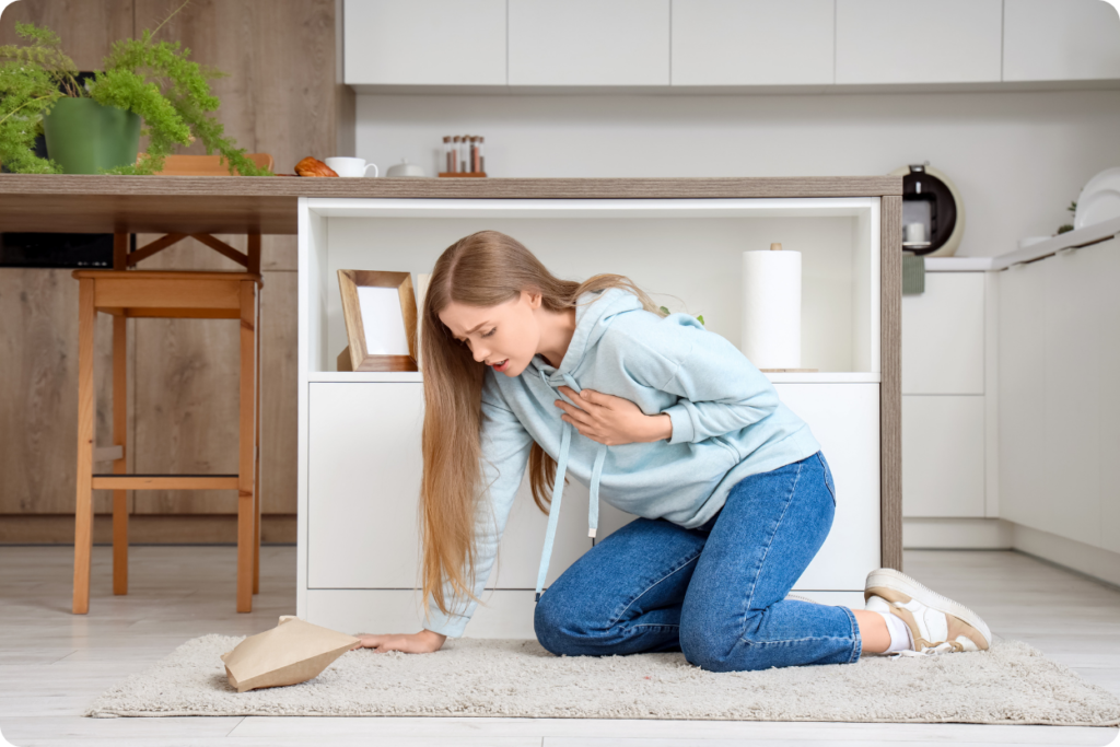 Mulher jovem de cabelos longos, vestindo moletom azul claro e calça jeans, ajoelhada no chão de uma cozinha enquanto pressiona o peito com uma expressão de dor e dificuldade para respirar.